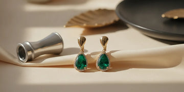 Pair of emerald earrings displayed on a neutral surface beside a jewelry loupe