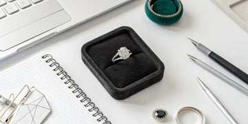 Custom engagement ring design in a box on a desk with a laptop and design notes.