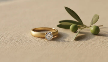 Minimalist gold solitaire engagement ring resting on linen with an olive leaf